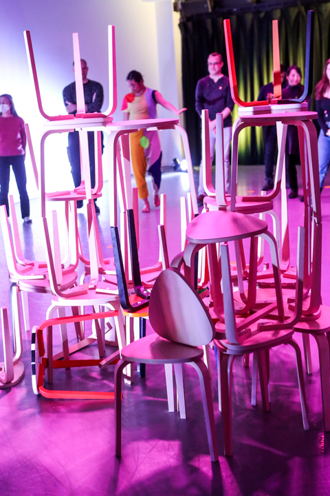 Pink three-legged stools piled one on the other; the top stools' legs are reaching ceiling ward. Dancers and audience members are in the background. A lilac color streams up one wall while a black curtain streaked with greenish light covers the other